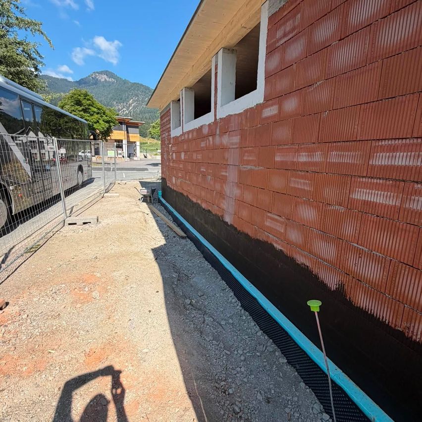 Eine Baustelle mit einem geparkten Bus an der Seite und einer Person, die ein Foto macht. Die Wand wird mit roten Ziegelsteinen gestrichen und hat ein blaues Entwässerungssystem.