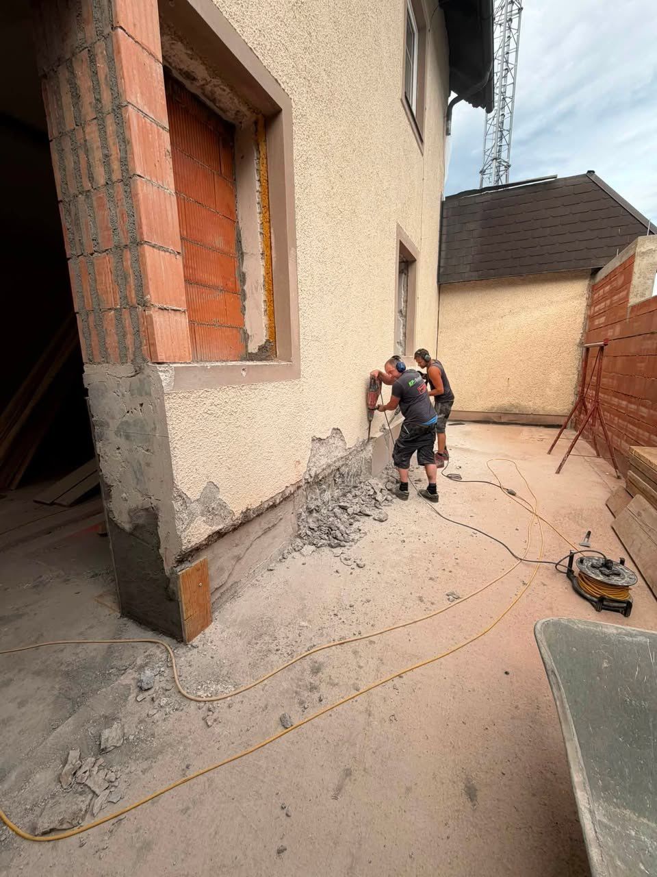 Zwei Männer arbeiten an einer Ziegelmauer, einer bohrt und der andere steht daneben. Ein Haufen Schutt liegt auf dem Boden, und ein Gebäude mit einem braunen Dach ist im Hintergrund.
