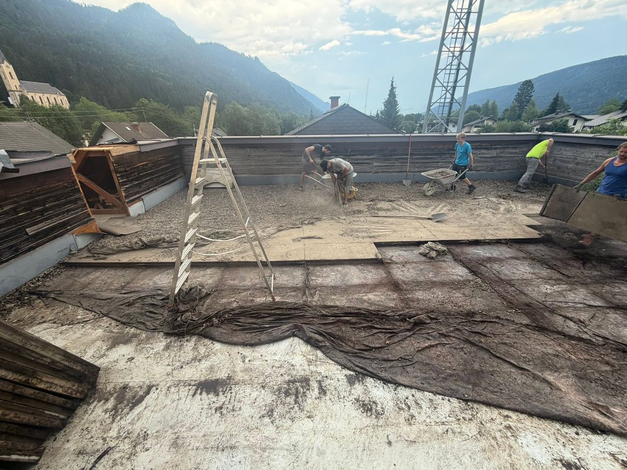 Several workers on a rooftop with mountains in the background, two are mixing cement while another pushes a wheelbarrow.