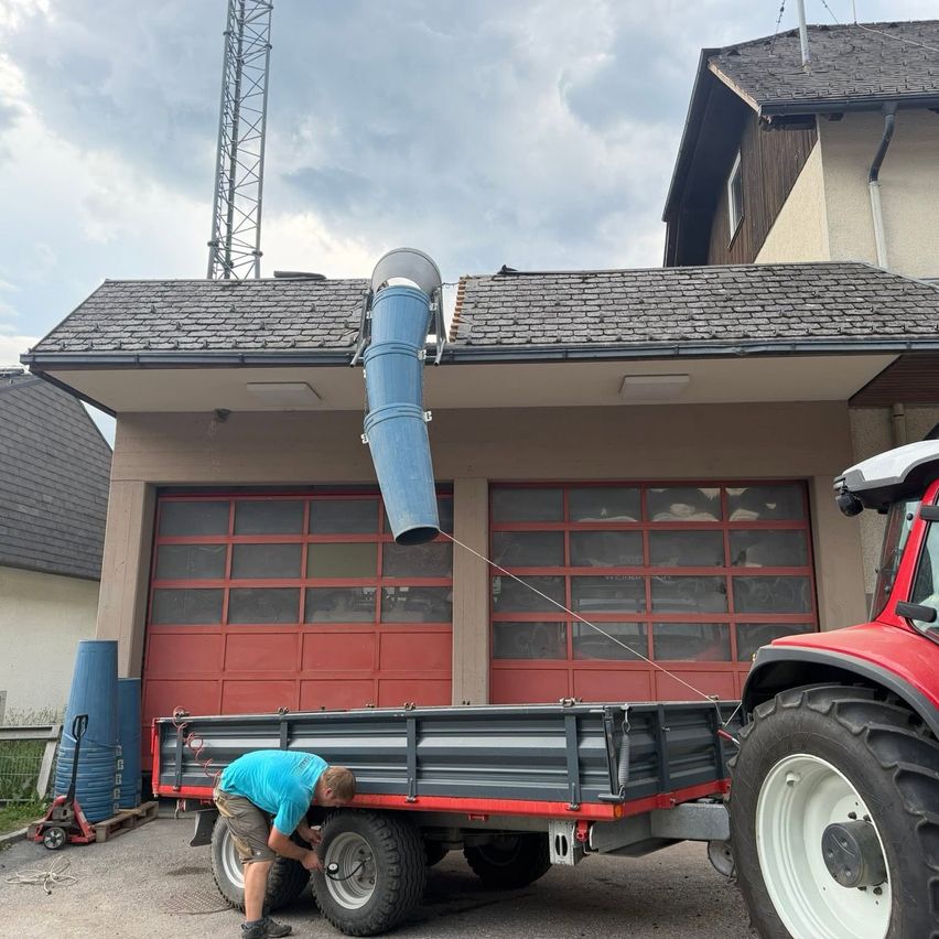 A man is working on a red tractor with a large blue container on the roof of a house with red garage doors.