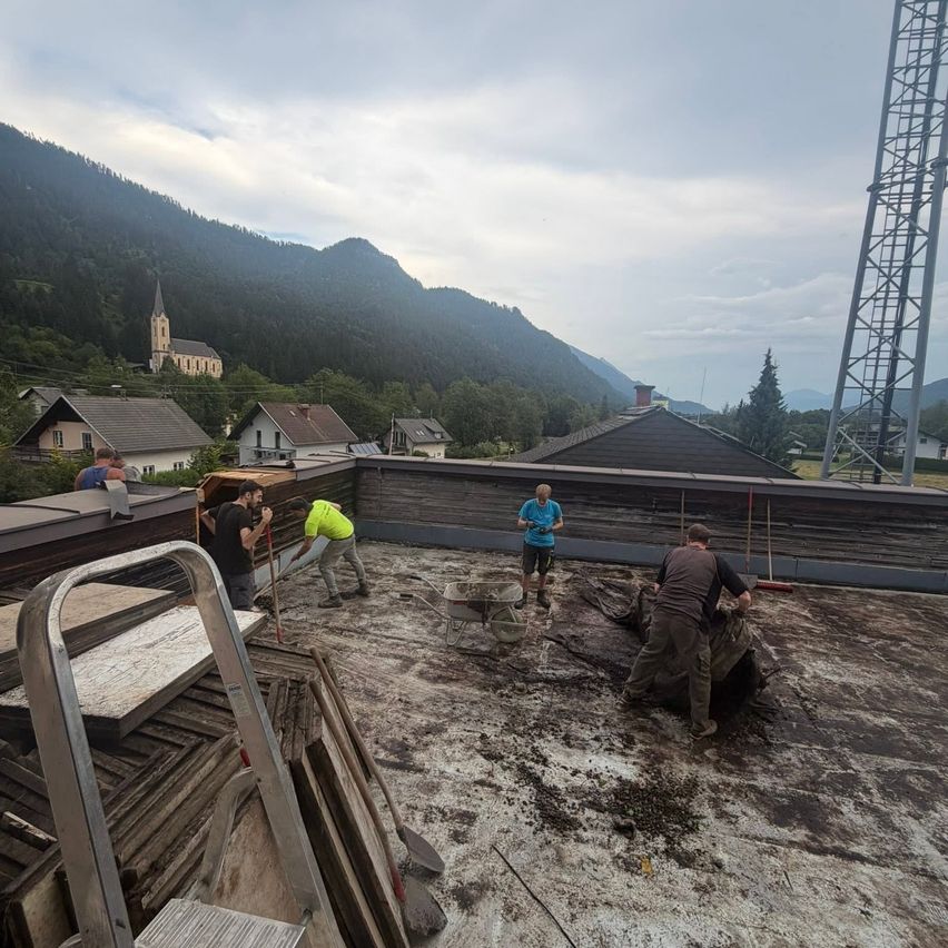 Five people are working on a rooftop, with one in a yellow shirt and two in black. They are using shovels and wheelbarrows, with mountains and a church in the background.