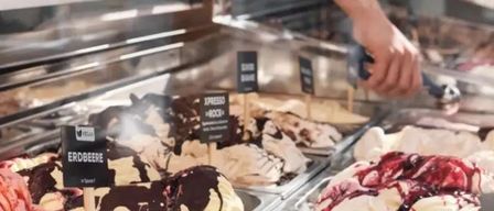 A person scoops ice cream from a display case, offering various flavors. The ice cream includes chocolate, strawberry, and mixed berry flavors. The display case is filled with different colored ice creams.