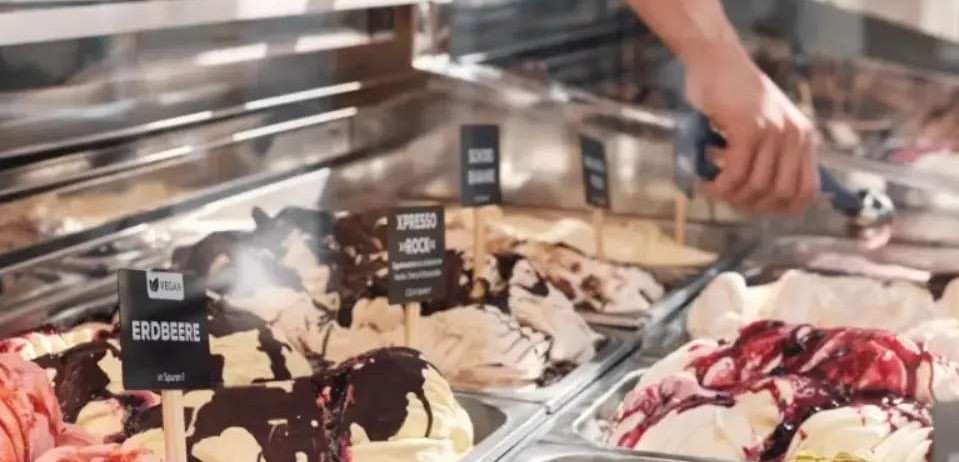 Eine Person holt Eis aus einer Vitrine und bietet verschiedene Geschmacksrichtungen an. Das Eis umfasst Schokolade, Erdbeere und gemischte Beeren. Die Vitrine ist mit Eis in verschiedenen Farben gefüllt.