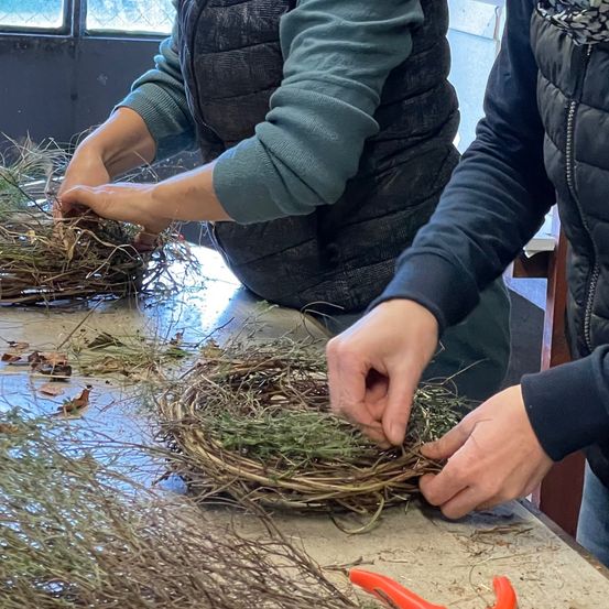Zwei Frauen arbeiten an einem Nest aus Zweigen und Gras, eine trägt eine Brille und die andere einen Schal.