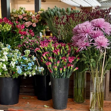 Ein Holztisch zeigt eine Vielzahl von Blumen in schwarzen Töpfen und durchsichtigen Vasen, darunter Rosen, Gänseblümchen und andere bunte Blüten.
