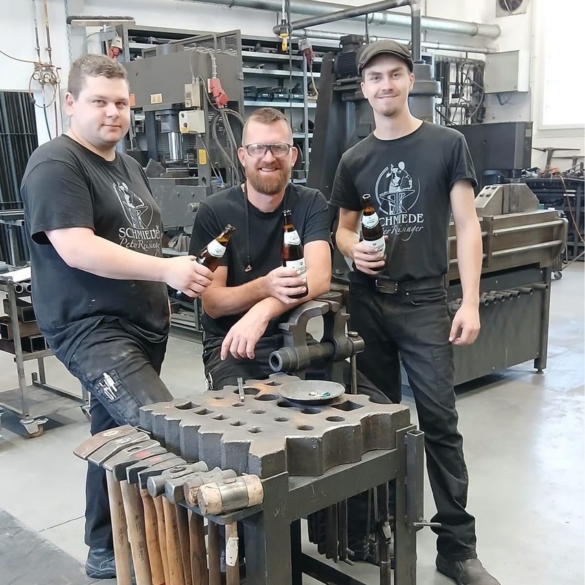 Drei Männer stehen in einer Werkstatt, jeder hält eine Bierflasche. Sie lächeln und posieren für ein Foto vor einer Metallbearbeitungsmaschine.