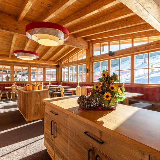 Ein Restaurant mit Holzbalken und Blick auf einen verschneiten Berg, mit Sonnenblumen, Holztischen und einer Theke mit Schubladen.