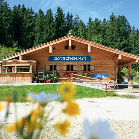 Eine Holzhütte namens Gotschenalm befindet sich in einem bewaldeten Gebiet mit einem grünen Rasen davor. Das Gebäude hat eine kleine Veranda mit einer Tafel und einigen Blumen.