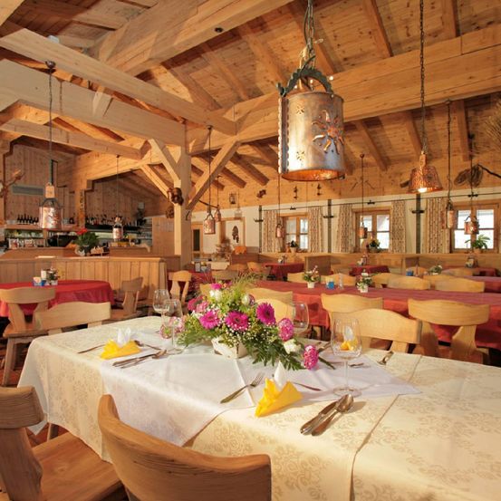 Ein Restaurant mit Holzbalken und einem großen Tisch, der für eine Mahlzeit gedeckt ist. Der Tisch hat einen Blumenstrauß in der Mitte, Weingläser und Besteck. Andere Tische sind aufgebaut, und der Raum ist warm beleuchtet mit Pendelleuchten.