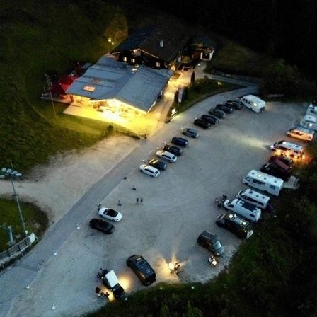 Eine Nachtaufnahme eines Parkplatzes mit mehreren geparkten Autos, einige mit eingeschalteten Scheinwerfern, vor einem beleuchteten Gebäude.