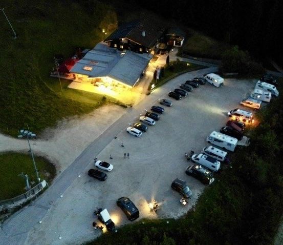 Eine Nachtaufnahme eines Parkplatzes mit mehreren geparkten Autos, einige mit eingeschalteten Scheinwerfern, vor einem beleuchteten Gebäude.