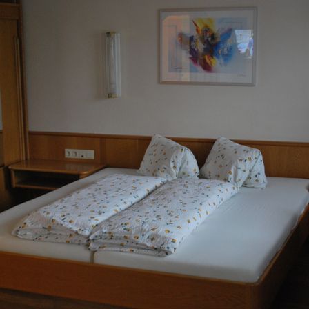 Ein Schlafzimmer mit einem Doppelbett, zwei Kissen und Bettwäsche mit Blumenmuster. Ein weißer Wandbilderrahmen und eine wandmontierte Leuchte. Ein Nachttisch mit Steckdosen und einem Holzkopfteil.