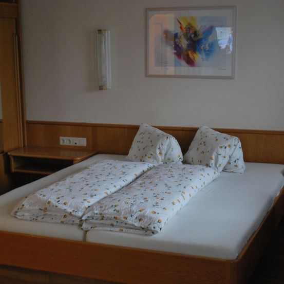 Ein Schlafzimmer mit einem Doppelbett, zwei Kissen und Bettwäsche mit Blumenmuster. Ein weißer Wandbilderrahmen und eine wandmontierte Leuchte. Ein Nachttisch mit Steckdosen und einem Holzkopfteil.