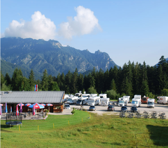 Ein Bergcampingplatz mit mehreren Wohnmobilen, die vor einem Gebäude mit Fahnen und Schirmen geparkt sind. Im Hintergrund befinden sich Bäume und Berge.