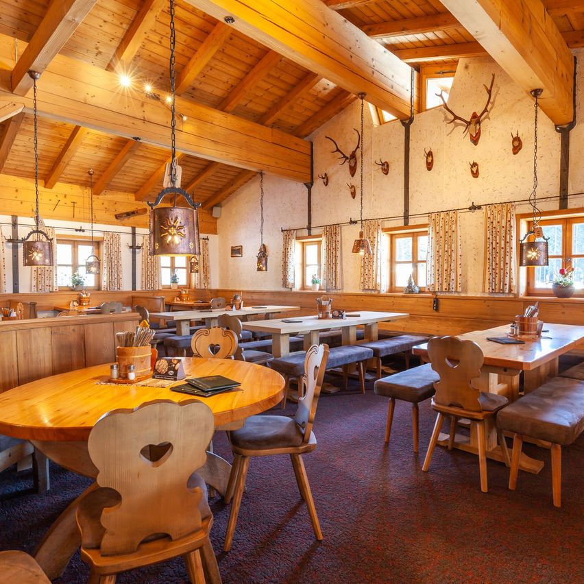 Ein rustikales Restaurant mit Holzbalken und Möbeln. Tische und Bänke sind aufgestellt, geschmückt mit dekorativen Gegenständen und Hirschgeweihen. Der Raum verfügt über große Fenster und Vorhänge, mit Lampen, die von der Decke hängen.