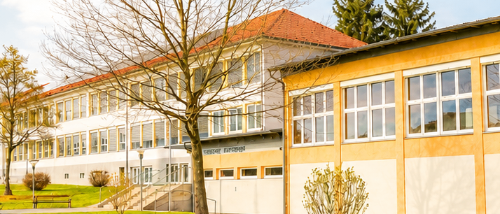 Ein modernes Schulgebäude mit rotem Dach und gelben Akzenten, umgeben von Bäumen und Gras. Das Gebäude hat viele Fenster und eine Treppe, die zum Eingang führt.