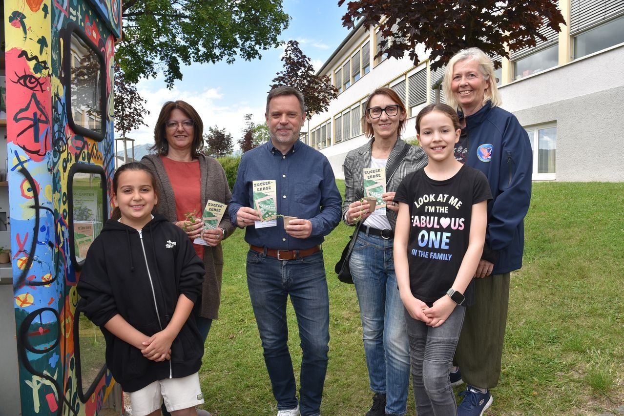 Eine Gruppe von Menschen, darunter Erwachsene und Kinder, steht vor einem Gebäude, lächelt und hält kleine Pflanzen. Der Mann in der Mitte hält ein Buch und eine Pflanze. Die Frau auf der rechten Seite trägt eine Uhr.