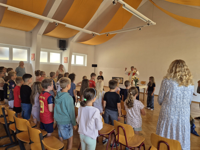 Eine Gruppe von Kindern und Erwachsenen steht in einem Raum, möglicherweise einer Kirche, während ein Priester eine Zeremonie leitet. Der Raum hat Holzstühle, einen Tisch mit Blumen und einen Lautsprecher.