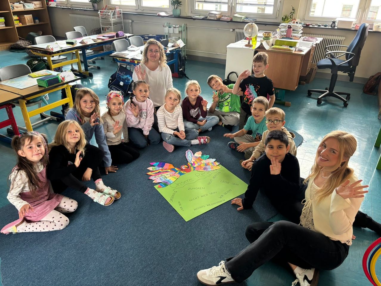 Eine Gruppe von Kindern und zwei Frauen sitzt im Kreis um ein farbenfrohes Papierbastelwerk auf dem Boden. Die Kinder lächeln und machen Handzeichen. Die Frau rechts lächelt und schaut in die Kamera. Der Klassenraum hat einen blauen Teppich, Schreibtische und Stühle.
