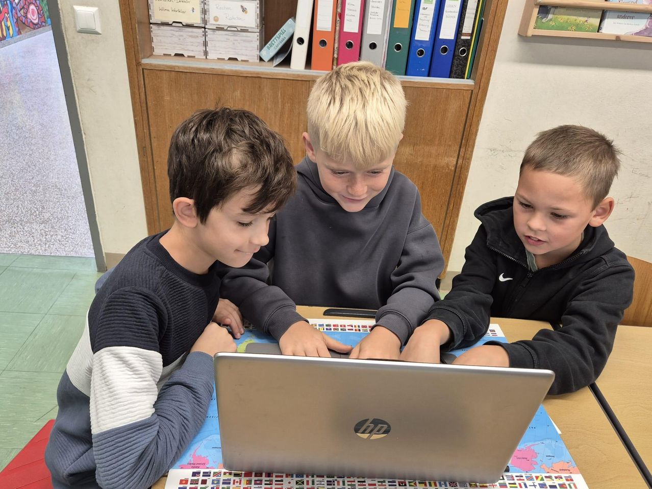Drei junge Jungen sitzen an einem Schreibtisch und schauen auf einen Laptop. Der Junge auf der linken Seite trägt einen grauen und weißen Pullover, der mittlere Junge trägt eine graue Kapuzenjacke und der Junge auf der rechten Seite trägt eine schwarze Kapuzenjacke. Der Laptop ist auf einer bunten Karte geöffnet und die Jungen konzentrieren sich auf den Bildschirm.