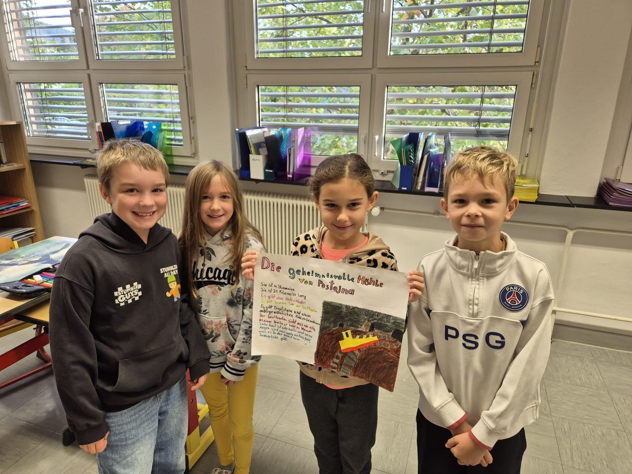 Vier Kinder in einem Klassenzimmer lächeln und posieren für ein Foto. Eines der Mädchen hält ein Poster mit einer Zeichnung eines Zuges und Text.