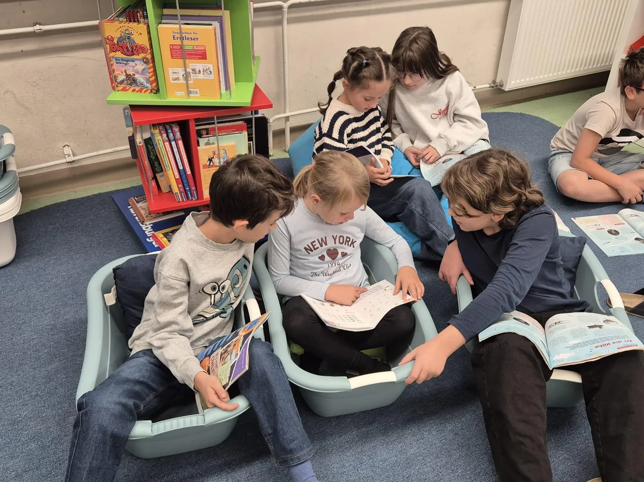 Eine Gruppe von Kindern sitzt auf Stühlen in einem Klassenzimmer. Einige lesen Bücher, während andere Bücher betrachten, die von anderen gehalten werden.