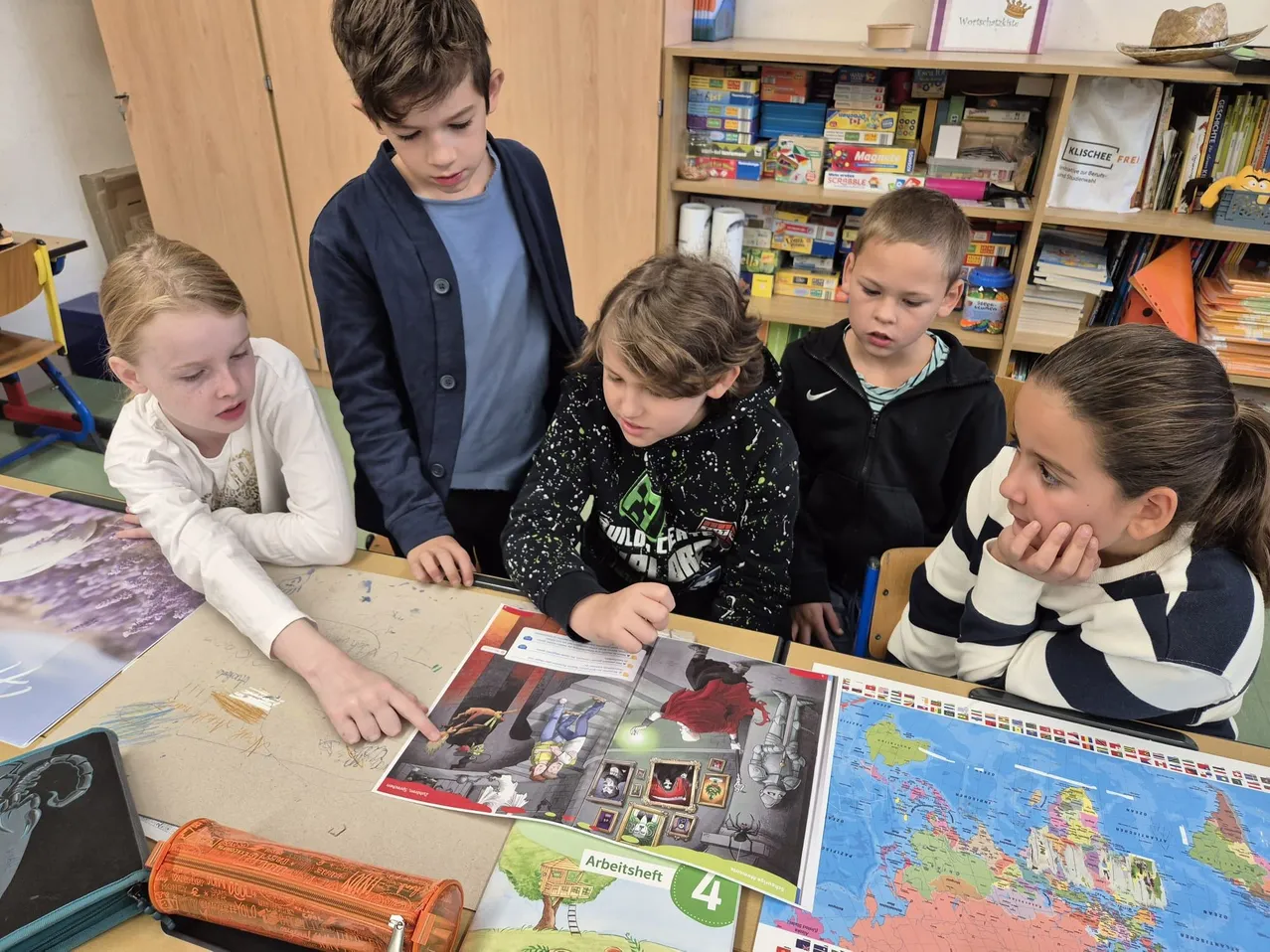 Fünf Kinder versammeln sich in einem Klassenzimmer um einen Tisch und betrachten ein Buch mit einer Karte und Bildern.