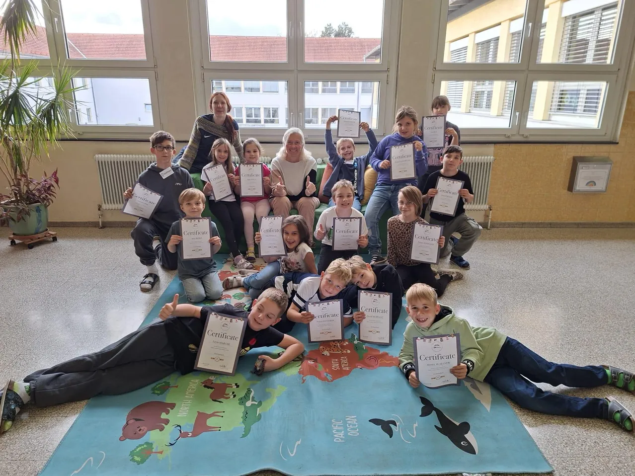 Eine Gruppe von Kindern und Erwachsenen posiert für ein Foto in einem Klassenzimmer. Sie halten Urkunden und sitzen auf einem Teppich mit einer Weltkarte.