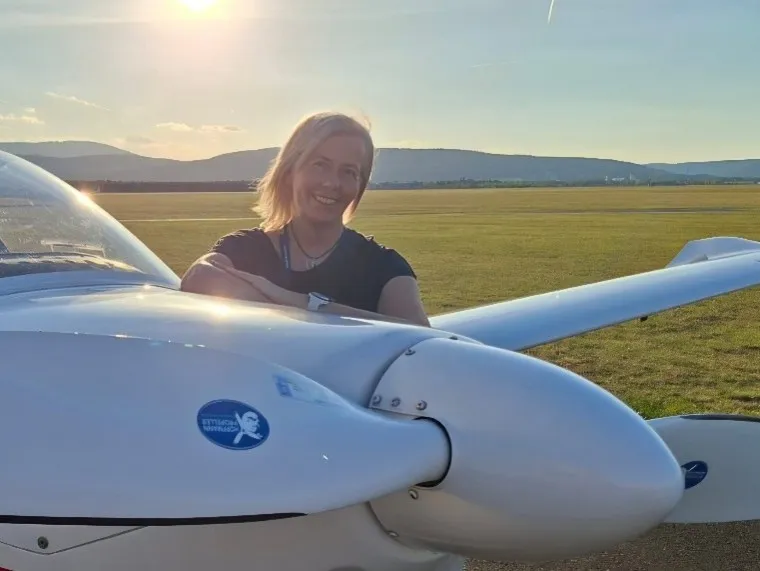 Eine Frau mit blonden Haaren lächelt und lehnt sich an die Vorderseite eines weißen Flugzeugs.