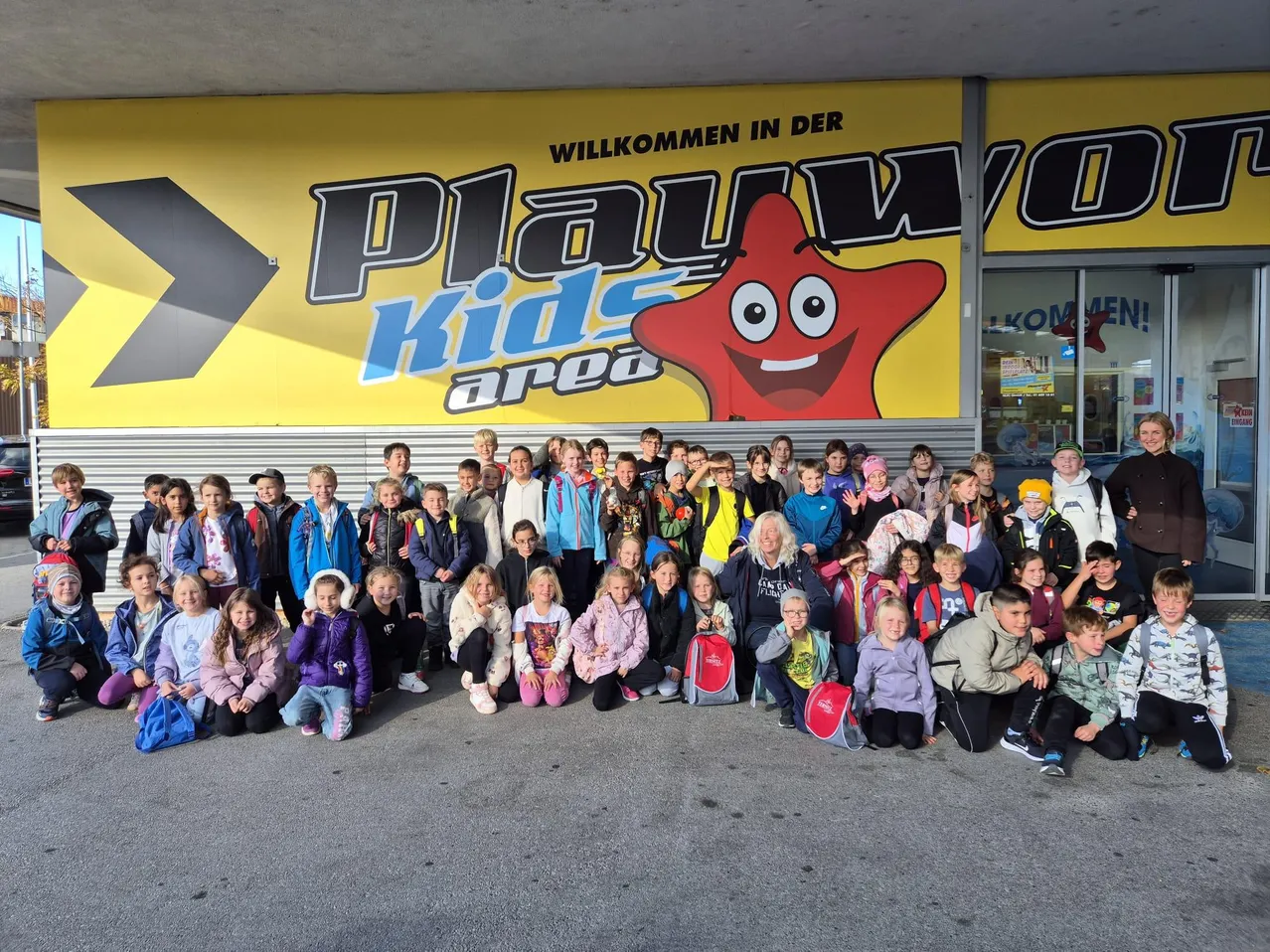 Eine Gruppe von Kindern und Erwachsenen posiert für ein Foto vor einem Gebäude mit einem Schild, das 'Playworld Kids Area' sagt.