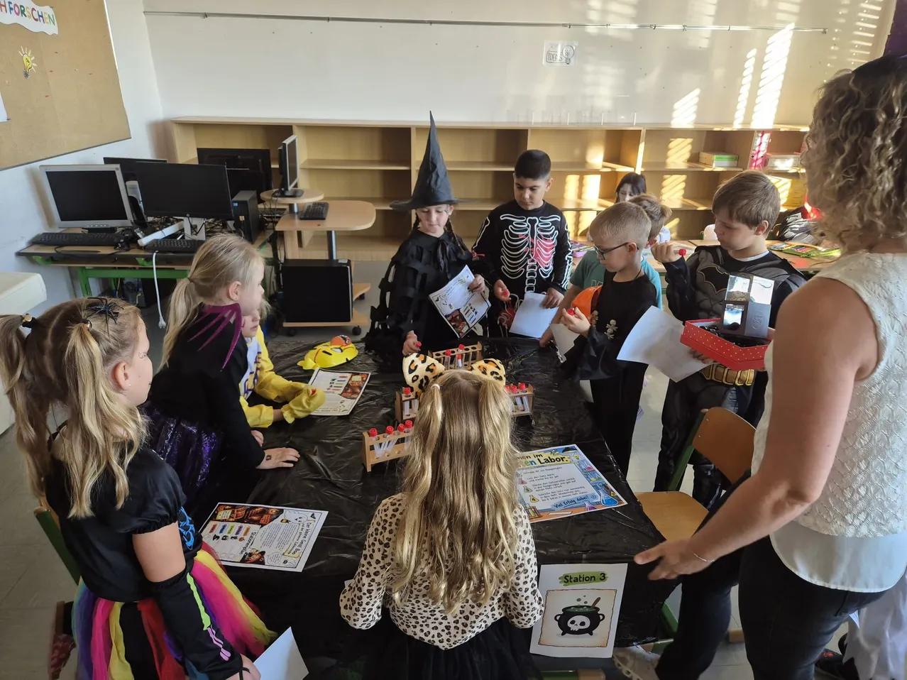 Eine Gruppe von Kindern versammelt sich in einem Klassenzimmer um einen Tisch, alle in Halloween-Kostümen gekleidet. Sie halten Papiere und betrachten einen Tisch mit einem Modell eines Labors. Eine Frau steht neben ihnen und hält ein Papier.