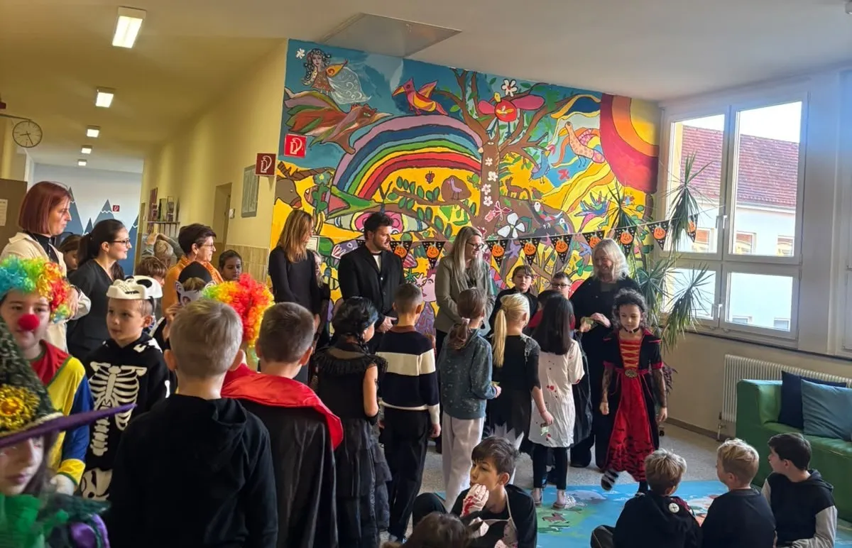 Eine Gruppe von Kindern und Erwachsenen versammelt sich in einem Raum. Die Kinder tragen Halloween-Kostüme. Der Raum hat ein farbenfrohes Wandbild an der Wand und es gibt Pflanzen und eine Uhr an der Wand.