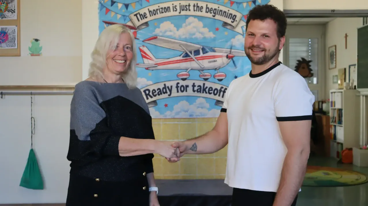 Eine Frau mit weißen Haaren und ein Mann mit Bart schütteln sich die Hände vor einem Banner, das 'Der Horizont ist nur der Anfang. Bereit für den Start.' sagt.