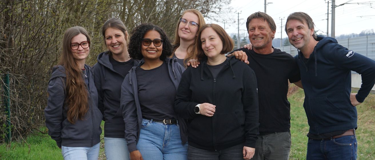 Sieben Menschen, sechs Frauen und ein Mann, stehen zusammen auf einem Kiesweg und lächeln für ein Foto. Sie tragen Freizeitkleidung, und der Hintergrund zeigt Grünflächen und Bäume.