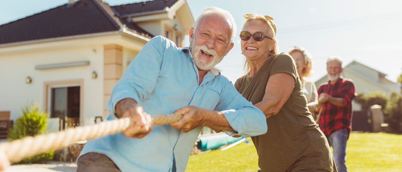 Eine Gruppe älterer Menschen hat Spaß im Garten eines Hauses. Ein Mann und eine Frau ziehen an einem Seil und lachen. Dahinter stehen andere, lächelnd und genießen den sonnigen Tag.