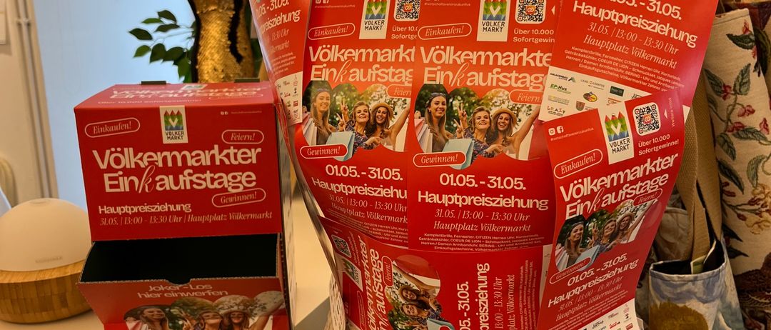 Eine Puppe mit Werbematerialien für die Veranstaltung Volksmarkt Ein/aufstage, ausgestellt auf einem Tisch mit Veranstaltungsboxen und einer Pflanze im Hintergrund.