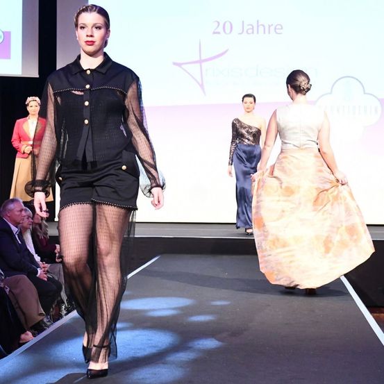 Eine Modenschau mit Models auf dem Laufsteg, das Publikum beobachtet. Eines der Models trägt ein schwarzes Outfit, die anderen tragen blaue und beige Kleider.