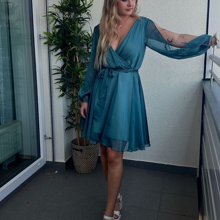 Eine Frau in einem türkisfarbenen Kleid mit durchsichtigen Ärmeln posiert auf einem Balkon mit einer Pflanze im Hintergrund.