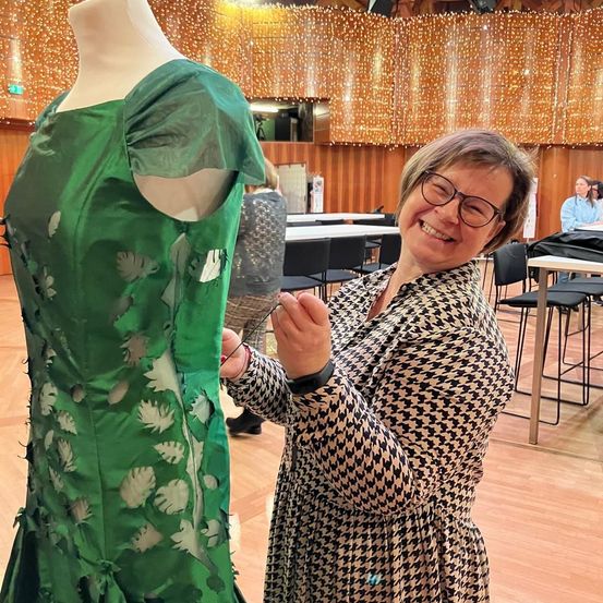 Eine Frau in einem schwarz-weißen Kleid mit einem Houndstooth-Muster hält eine Nadel und Faden und repariert ein grünes Kleid an einem Mannequin. Im Hintergrund stehen Tische und Stühle, und eine Frau in einem blauen Shirt steht.