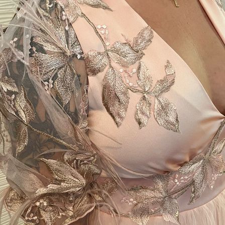 Eine Nahaufnahme eines rosa Abendkleides mit Goldblattstickerei, zarten Spitzenärmeln und einem transparenten Oberteil. Das Design zeigt ein Muster aus Blättern und Blumen, was ihm einen eleganten und festlichen Look verleiht.