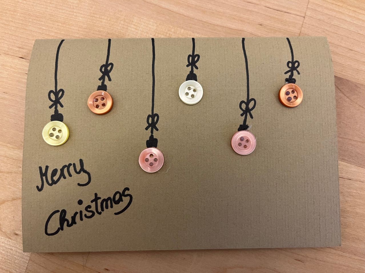 Eine handgefertigte Weihnachtskarte mit 'Merry Christmas' in schwarzer Tinte geschrieben. Sie zeigt sechs kupferfarbene Knöpfe, die an schwarzen Fäden von einem braunen Hintergrund hängen.