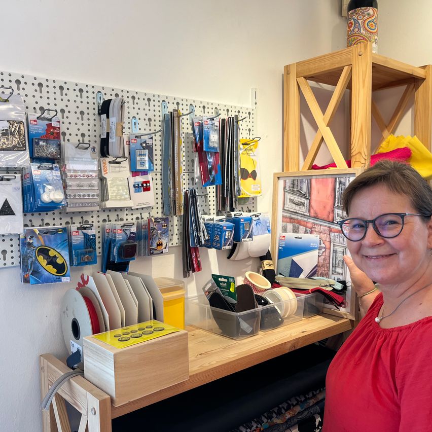 A woman stands in a craft room filled with various sewing supplies, including Batman-themed items, arranged on a pegboard and a wooden desk.
