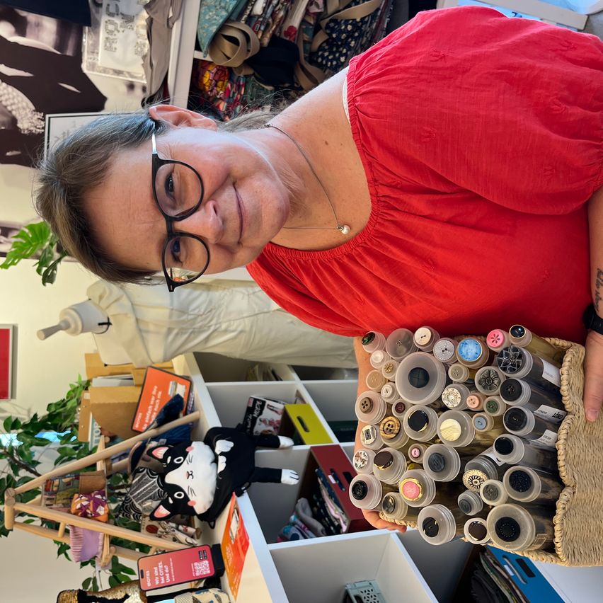 A woman in a red shirt and glasses holds a basket filled with various buttons. She is standing in a room with a white shelf and a stuffed cat in the background.