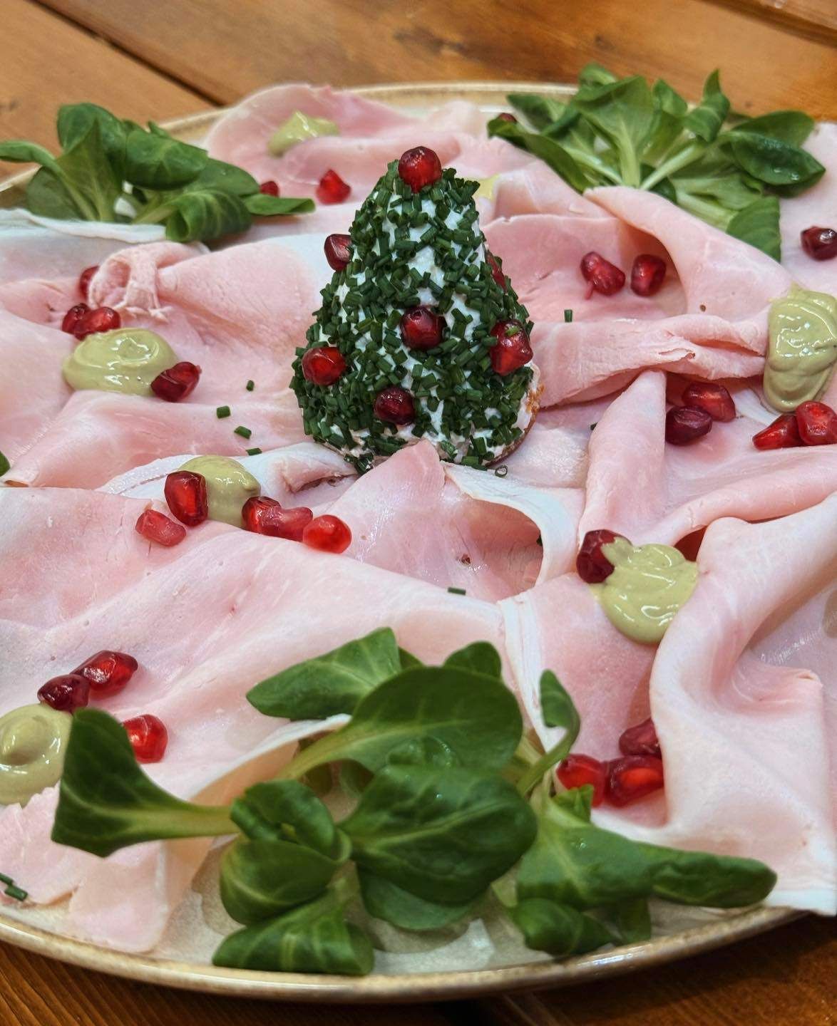 Ein Gericht aus dünn geschnittenem rosa Fleisch, vielleicht Schinken, ist mit grünen Kräutern und roten Granatapfelsamen arrangiert. Eine baumförmige Käsegarnitur befindet sich in der Mitte.