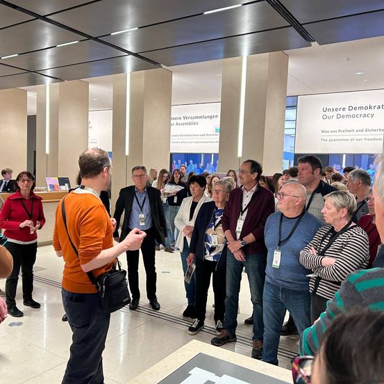 Eine Gruppe von Menschen steht in einem Saal und hört einem Mann zu, der vor ihnen spricht. Einige Leute tragen Namensschilder. Der Saal hat eine hohe Decke mit Lichtern und einer großen Leinwand mit Text.