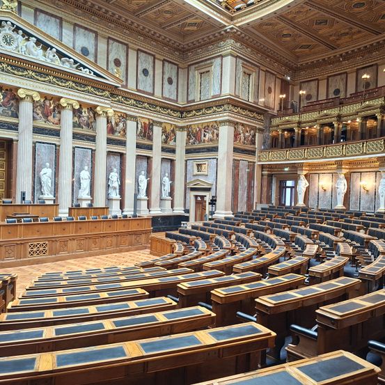 Ein leerer Parlamentssaal mit Holzbänken, Marmorsäulen und Statuen. Die Decke ist kunstvoll mit goldenen Designs.