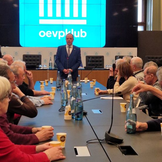 Eine Versammlung mit einem älteren Publikum, ein Mann in Anzug steht an einem Rednerpult mit Mikrofon, und das Publikum sitzt an einem langen Tisch mit Tassen und Flaschen.