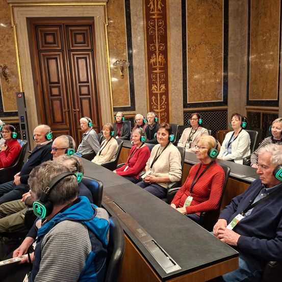Eine Gruppe von Menschen mit grünen Kopfhörern sitzt in einem Konferenzraum in Stühlen. Sie schauen alle nach vorne, wahrscheinlich einer Präsentation zuhörend. Der Raum hat eine Marmorwand und eine Holztür.