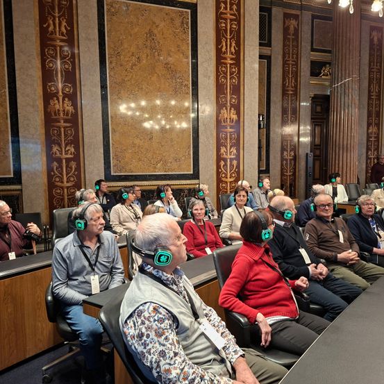 Eine Gruppe älterer Erwachsener mit Kopfhörern mit grünen Lichtern sitzt in einem Konferenzraum mit verzierten Wänden und einer Kristallleuchte.
