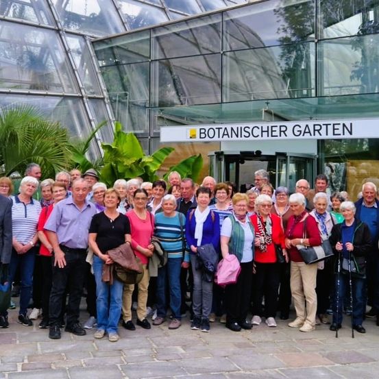 Eine Gruppe von Menschen in lässiger Kleidung steht vor einem Glasgebäude mit dem Schild 'Botanischer Garten' über dem Eingang.